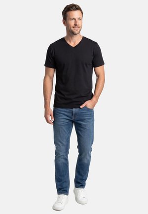 Mann mit kurzen Haaren, der ein schwarzes V-Ausschnitt-T-Shirt, blaue Jeans und weiße Sneakers trägt, steht mit einer Hand in der Tasche vor weißem Hintergrund.