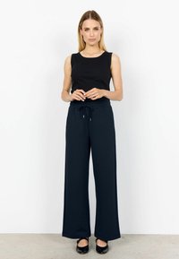 Woman with blonde hair wears black sleeveless top, navy wide-leg drawstring pants, and black flats, standing against a white wall.
