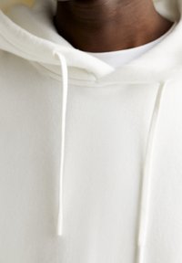 Close-up van een persoon die een eenvoudige witte hoodie draagt met zichtbare trekkoorden en een wit innerlijk shirt onder de halslijn.