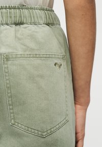 Κοντινό πλάνο σε παντελόνι με ελαστική μέση σε ανοιχτό πράσινο χρώμα, με πίσω τσέπη που έχει μικρό λογότυπο σε σχήμα καρδιάς στη δεξιά πλευρά.