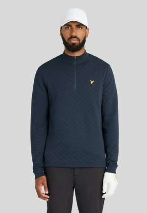 Mand med skæg iført hvid kasket, sort quiltet langærmet poloshirt med gul logo, sorte bukser og hvid golfhandske på højre hånd.