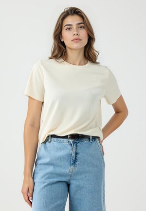 T-shirt crème à manches courtes avec un col rond et une texture lisse, associé à un jean en denim bleu taille haute et une ceinture noire.