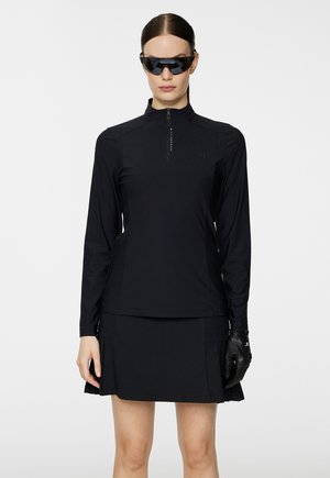 Black long-sleeve top with zipper detail, paired with a black pleated skirt. Model wears black sunglasses and leather gloves, minimalistic design.