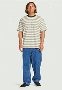 Homem de pé, vestindo uma t-shirt às riscas bege e verde, jeans azul largos e ténis pretos, em fundo claro.