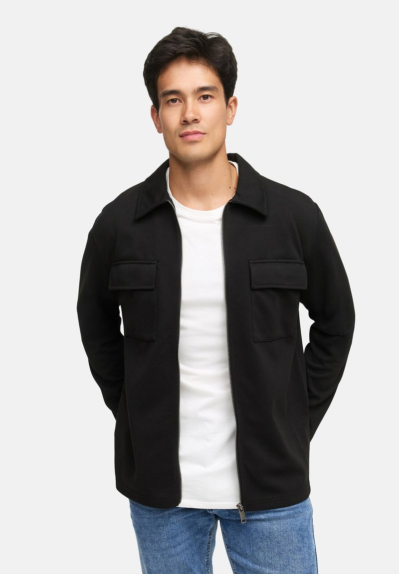 Junger Mann mit dunklem Haar, der eine schwarze Jacke mit Reißverschluss über einem weißen Hemd und blauen Jeans trägt und mit den Händen hinter dem Rücken steht.