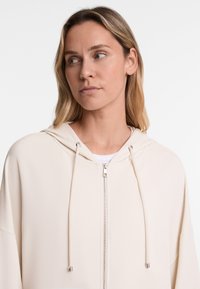Svijetla bež zip-up dukserica s kapuljačom na vezanje, ležernog kroja i glatkog materijala. Sadrži srebrni patentni zatvarač i detalje.