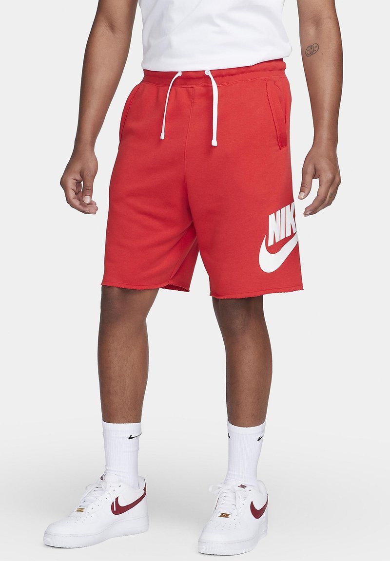 Personne portant un short rouge Nike avec un cordon blanc, des chaussettes blanches Nike et des baskets blanches avec un logo Swoosh rouge, debout devant un fond clair.