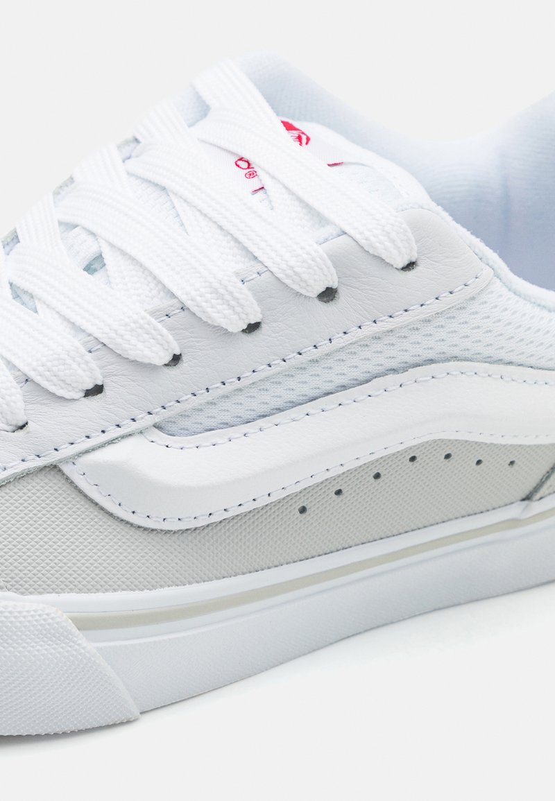 Zapatilla blanca con parte superior de malla y cuero, que presenta costuras tonales, cordones planos, suela de goma y acentos perforados en el lateral.