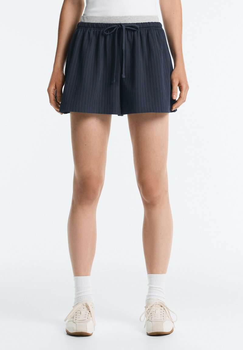 Personne portant un short bleu marine foncé à fines rayures avec une taille à cordon, des chaussettes blanches et des baskets couleur crème, debout sur un fond blanc.