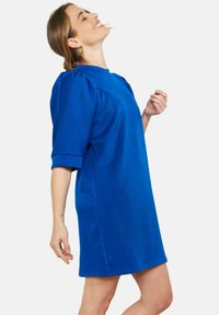 Frau trägt ein knielanges, hellblaues Kleid mit Puffärmeln, lächelt und legt den Kopf auf einem schlichten Hintergrund nach hinten.