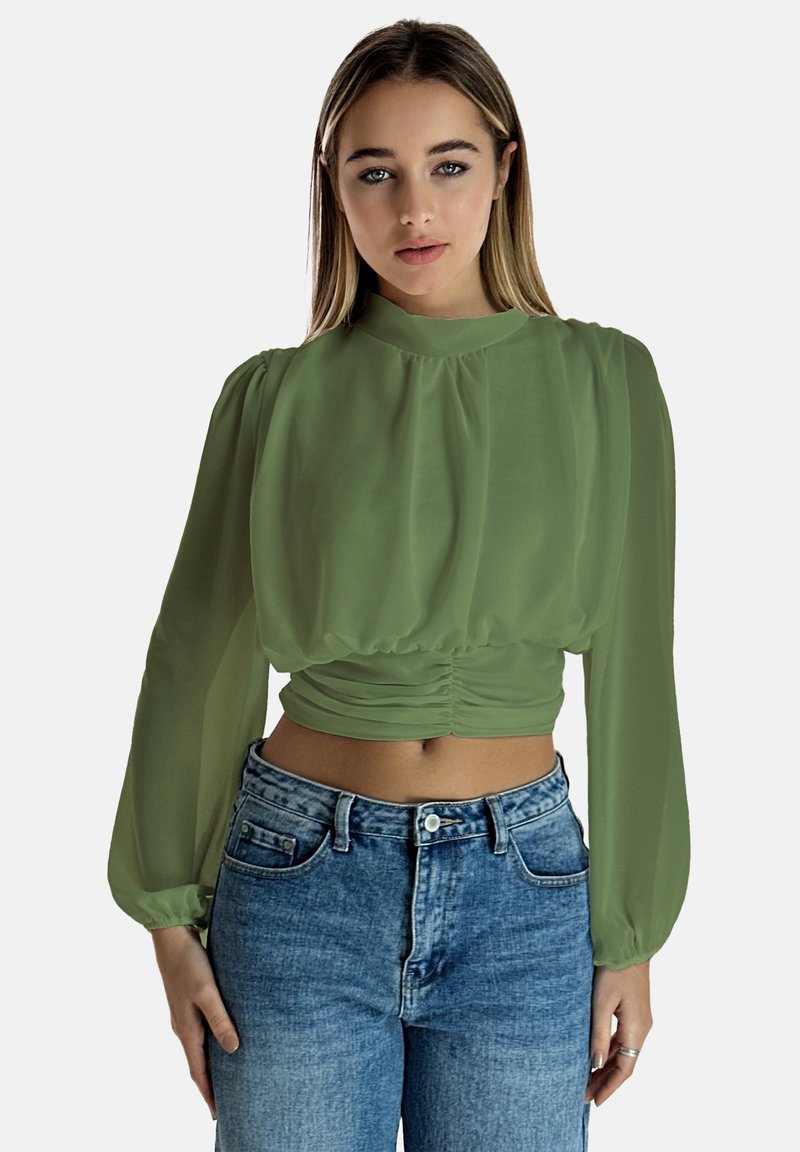 Blusa in chiffon verde con colletto alto, maniche lunghe plissè e vita arricciata, abbinata a jeans in denim a vita alta.