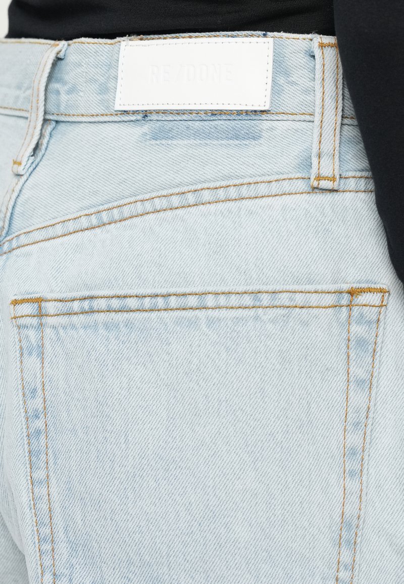 Lichtblauwe denim jeans met een medium taille, voorzien van een witte leren label met de tekst "RE/DONE" en contrasterende oranje stiksels.