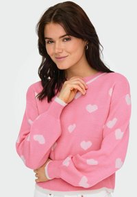 Roza pletena jopica z belimi srčastimi vzorci, okroglo izrezana in z rebrastimi manšeti. Tkanina se zdi mehka in teksturirana.