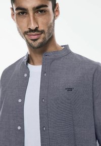 Camicia grigia con bottoni e un motivo testurizzato, dotata di colletto alla coreana e un logo sul petto. Indossata sopra una maglietta bianca.