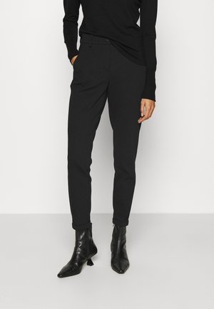 Persona que lleva pantalones negros ajustados con puños y botines negros de punta puntiaguda, de pie con una mano en el bolsillo contra un fondo blanco.