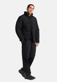 Schwarze Steppjacke mit hohem Kragen und seitlichem Reißverschluss, kombiniert mit locker sitzender schwarzer Cargohose und schwarzen Turnschuhen.