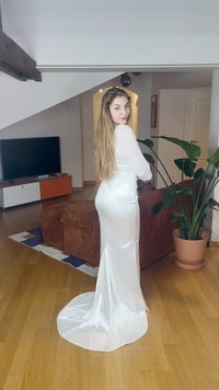 Hosszú hajú nő fehér selyem ruhában, aki egy fa padlós nappaliban áll, háttérben növény, kanapé és TV.