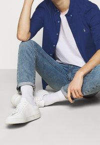 Marinblå skjorta med knappar över vit t-shirt, ljusblå slitna jeans, vita sneakers, sittande pose med korslagda ben och hand på hakan.