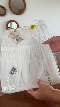 Hände, die eine gefaltete weiße Bluse in klarer Plastikverpackung mit Größenetiketten und Preisschildern halten, runde Holzspiegel an der Hintergrundwand.