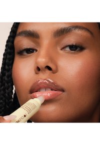 Un tubo di balsamo per labbra in mano, applicando una formula lucida e rosa chiaro sulle labbra. Presenta toni di pelle naturali e caldi e capelli intrecciati.