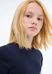 Top a coste rib a righe di colore blu marino con scollatura tonda. Il modello ha capelli biondi di lunghezza media e trucco minimal, con uno sfondo neutro.