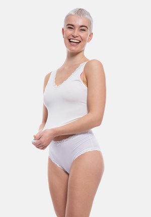 Persona sorridente con capelli corti che indossa una canottiera bianca con bordi in pizzo e biancheria intima abbinata su sfondo neutro.