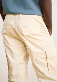 Beige cargo pants with multiple pockets, made of textured fabric. Features side and back pockets, with a fitted waist and reinforced seams.
