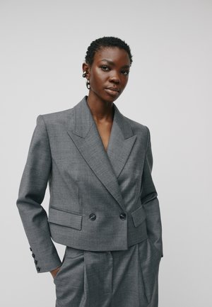 Femme avec des cheveux courts et bouclés portant un blazer gris ajusté à double boutonnage et un pantalon assorti, se tenant les mains dans les poches devant un fond blanc.
