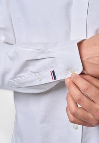 Vit långärmad skjorta med knäppbar manschett, prydd med en dekorativ randig detalj i blått, rött och vitt nära manschetten. Slätt, bomullsmaterial.