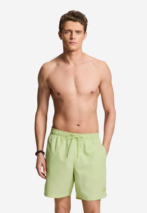 Ung mand med kort, krøllet hår iført lysegrønne badeshorts med snøre, står med den ene hånd i lommen mod en ensfarvet baggrund.