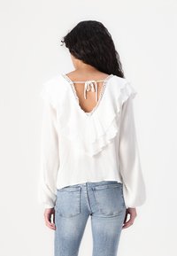 Camicia bianca a maniche lunghe con dettagli arricciati sulla schiena, colletto a V profonda e chiusura a laccio. Tessuto morbido e strutturato.