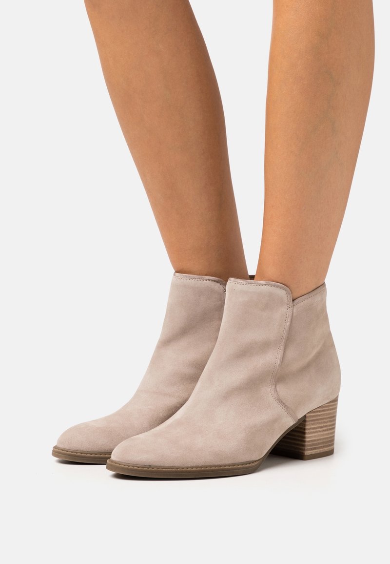 Bottes en daim beige montantes à la cheville avec talons blocs en bois empilés, portées sur jambes nues sur fond blanc.