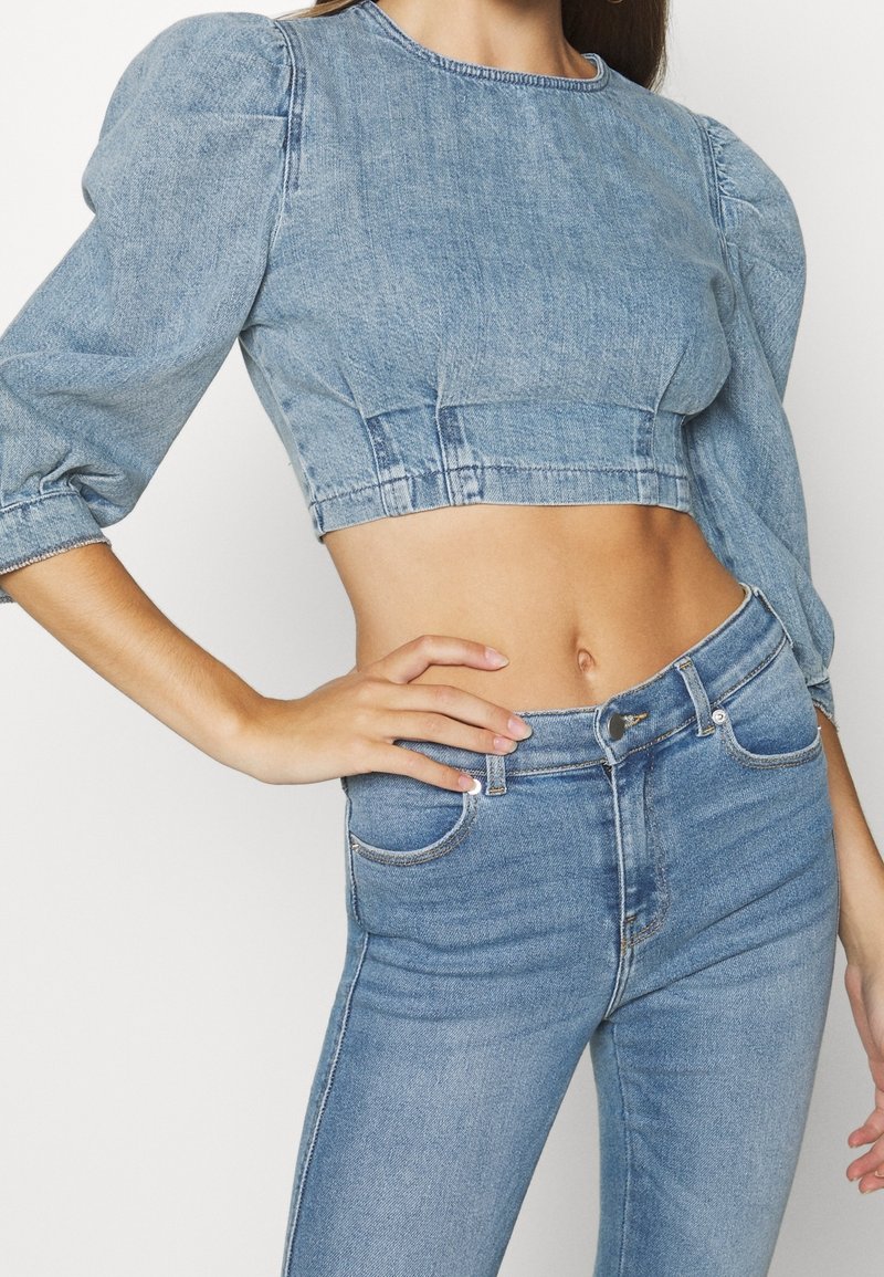 Femme portant un crop top en denim bleu clair avec des manches bouffantes et un jean taille haute en denim bleu, la main posée sur la hanche.