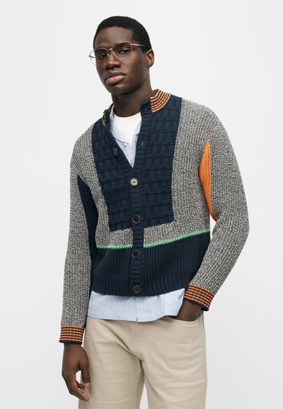 Άνδρας με γυαλιά, πολύχρωμο πλεκτό ζακέτα με κουμπιά, γαλάζιο πουκάμισο και μπεζ παντελόνι, όρθιος με το ένα χέρι στην τσέπη.
