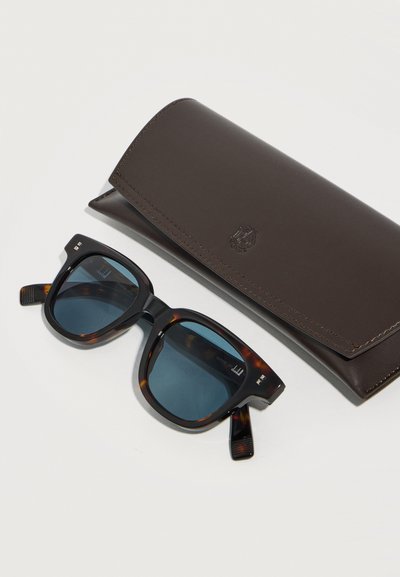 Μαύρα και καφέ τετράγωνα γυαλιά ηλίου με σκούρους φακούς δίπλα σε θήκη προστασίας από σκούρο καφέ δερματίνη με ανάγλυφο σχέδιο, πάνω σε λευκή επιφάνεια.