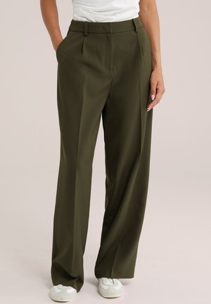 Personne portant un pantalon ample taille haute vert olive avec des poches et des baskets blanches, debout devant un fond uni.