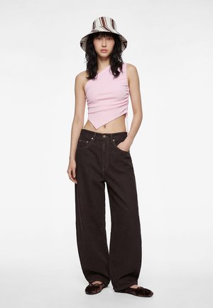 Femme portant un chapeau seau rayé, un crop top asymétrique rose, un pantalon large foncé et des ballerines en velours foncé, debout les mains dans les poches.