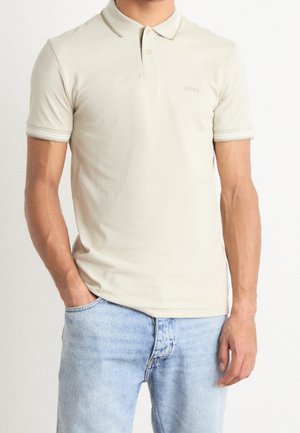 Mann trägt ein beiges kurzärmeliges Poloshirt mit Kragen und hellblaue Jeans, steht mit einer Hand in der Tasche.