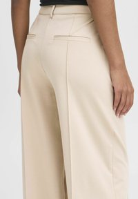 Beige Maßhose mit hoher Taille, ausgestattet mit zwei Gesäßtaschen und einem glatten, leicht strukturierten Stoff. Schlichte, gerade Beinform.