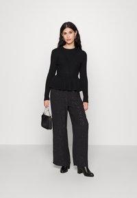Fekete bordázott peplum pulóver, széles lábú fekete nadrággal, amely finom csillogó mintával rendelkezik. A modell egy kis fekete táskát tart.