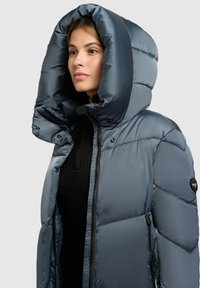 Šedá prešívaná bunda s veľkou nadýchanou kapucňou. Má zipsové zapínanie, bočné vrecká a pod ňou je textúrovaný čierny sveter.