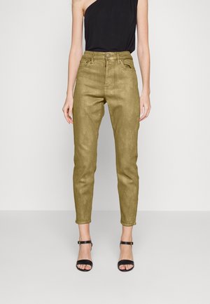 Žena nosí olivově zelené slim fit kalhoty s vysokým pasem, černý top na jedno rameno a černé otevřené boty na podpatku s páskem kolem kotníku, stojící proti bílému pozadí.