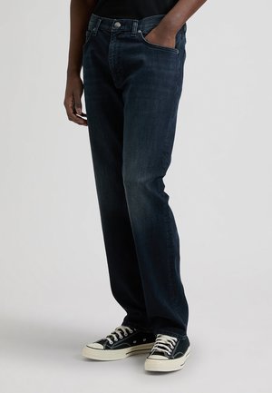 Person, die dunkelblaue Jeans und schwarze Sneaker mit weißen Sohlen trägt, steht mit einer Hand in der Tasche vor einem schlichten Hintergrund.