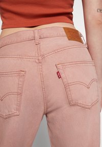 Jean en denim rose clair avec une coupe classique, comprenant deux poches arrière, des coutures orange et une  étiquette en cuir brun sur la ceinture.