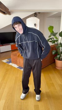 Tamsiai mėlynas su užtrauktuku džemperis su baltais siūlais, derinamas su juodomis kelnėmis ir sidabrinėmis sportinėmis avalynėmis. Vidaus interjeras su medinėmis grindimis ir augalais.