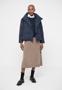 Marineblauw pufferjack met een cropped ontwerp, gecombineerd met een marineblauwe trui en een beige gebreide midi-rok. Zwarte loafers met gouden accenten maken de look compleet.