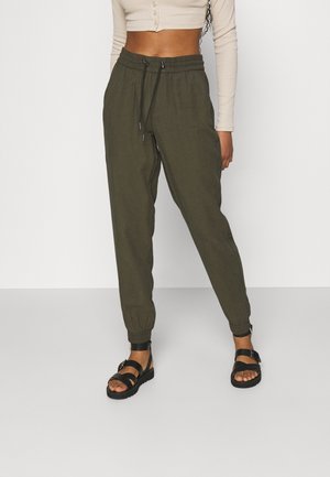 Olijfgroene, lichte joggingsbroek met elastische boorden en een trekkoord in de taille. Combinatie met zwarte sandalen met bandjes en een aansluitend beige top.
