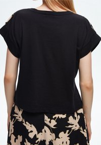 Svart kortärmad topp med avslappnad passform, med utskärningar på axlarna, tillverkad av mjukt tyg.