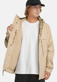 Beige vattentät jacka med en dragkedjeficka, framknappar, elastiska ärmslut och huva, bärs över en vit t-shirt.