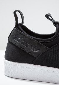 Černá teniska s plátěným vnějškem, gumovou podrážkou, patní částí z umělé kůže s vyraženým logem a poutkem pro snadné nazutí.